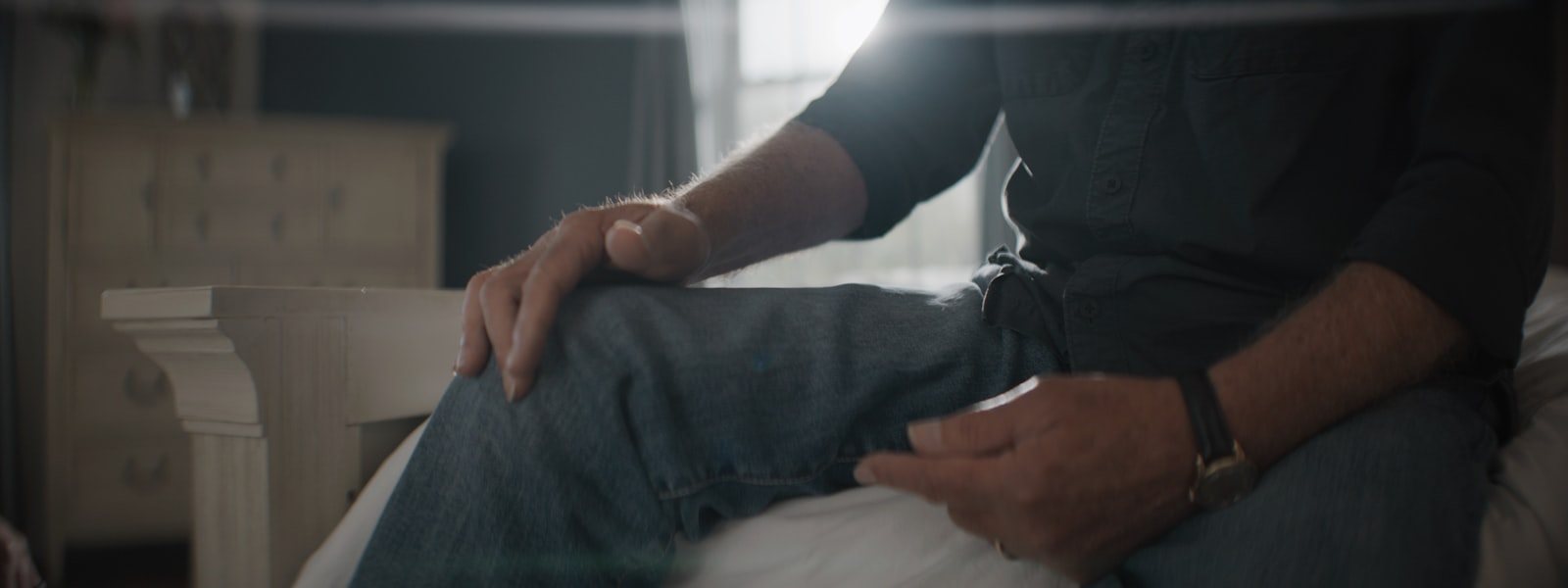 A person sitting on the edge of a bed with hands on their knees